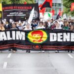 Eine Demonstration auf der Straße. Vorne ist ein großes Transparent mit der Aufschrift "Halim Dener" und dem Gesicht von Halim Dener. Im Hintergrund ist ein Transparent mit der Aufschrift "Von Rojava bis nach Berlin - Kommunales Leben aufbauen und verteidigen". Einige Menschen tragen das Gesicht von Halim Dener als Maske.