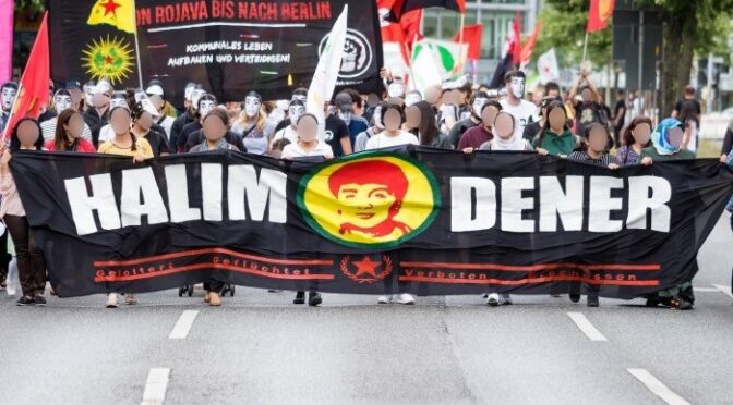 Eine Demonstration auf der Straße. Vorne ist ein großes Transparent mit der Aufschrift "Halim Dener" und dem Gesicht von Halim Dener. Im Hintergrund ist ein Transparent mit der Aufschrift "Von Rojava bis nach Berlin - Kommunales Leben aufbauen und verteidigen". Einige Menschen tragen das Gesicht von Halim Dener als Maske.